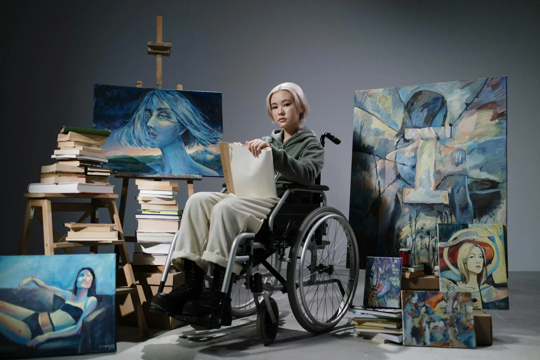 a woman in gray sweater sitting on wheelchair surrounded with books and paintings